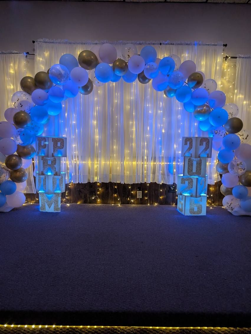 Blue balloon arch and white curtain backdrop with string lights, flanked by lit cube stands in an event space