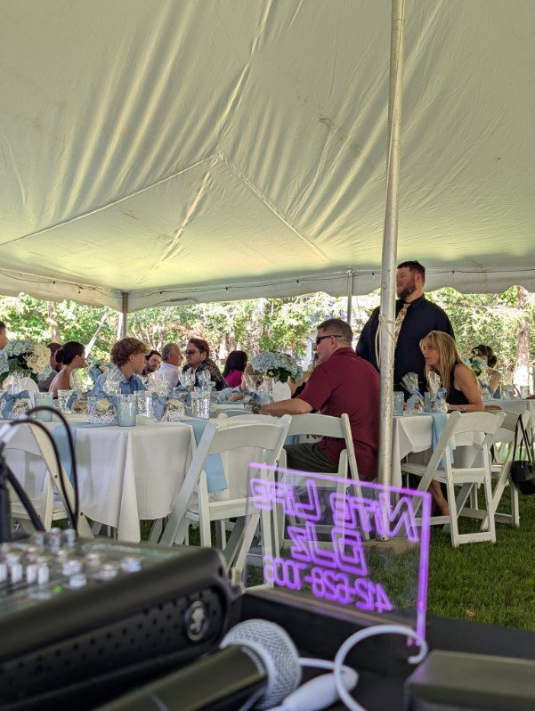 Guests seated at white-clothed tables under a tent at an outdoor event, with a glowing purple sign in front.