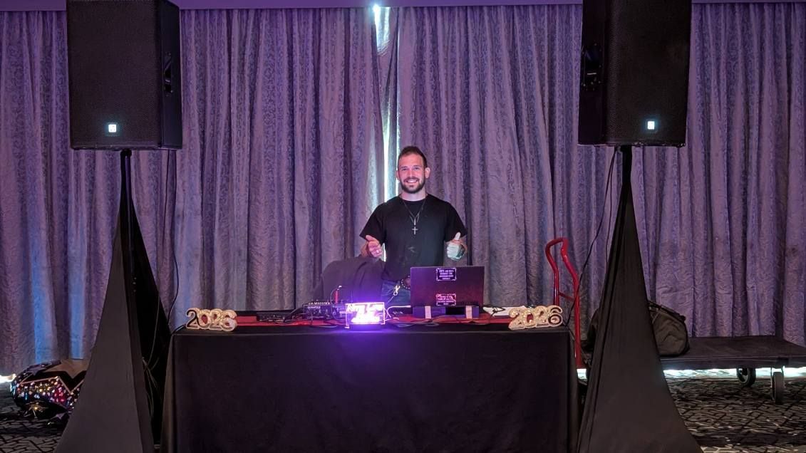 DJ standing behind a lit booth with purple curtains and speakers in the background