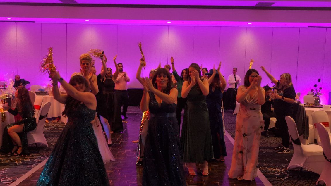Guests dancing at a purple-lit formal event with tables and floral centerpieces in the background