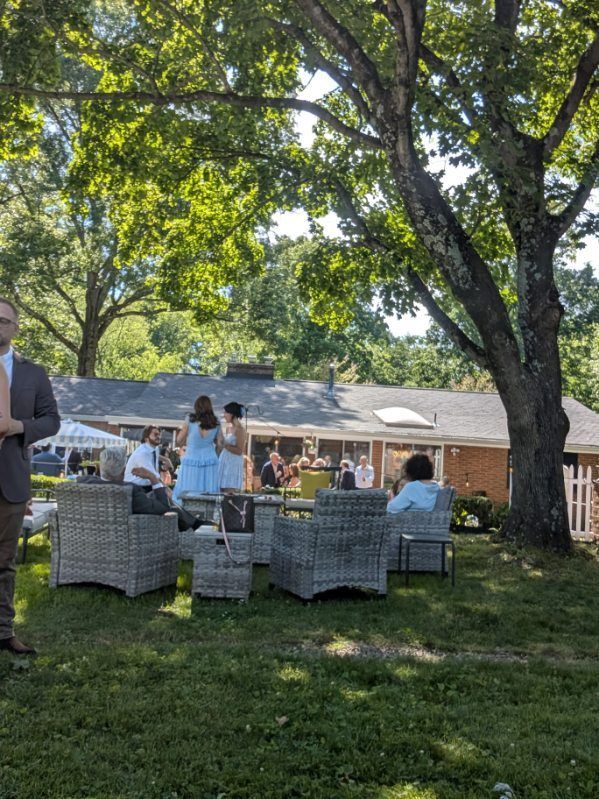 People seated on patio chairs under large trees in a sunny outdoor gathering area.