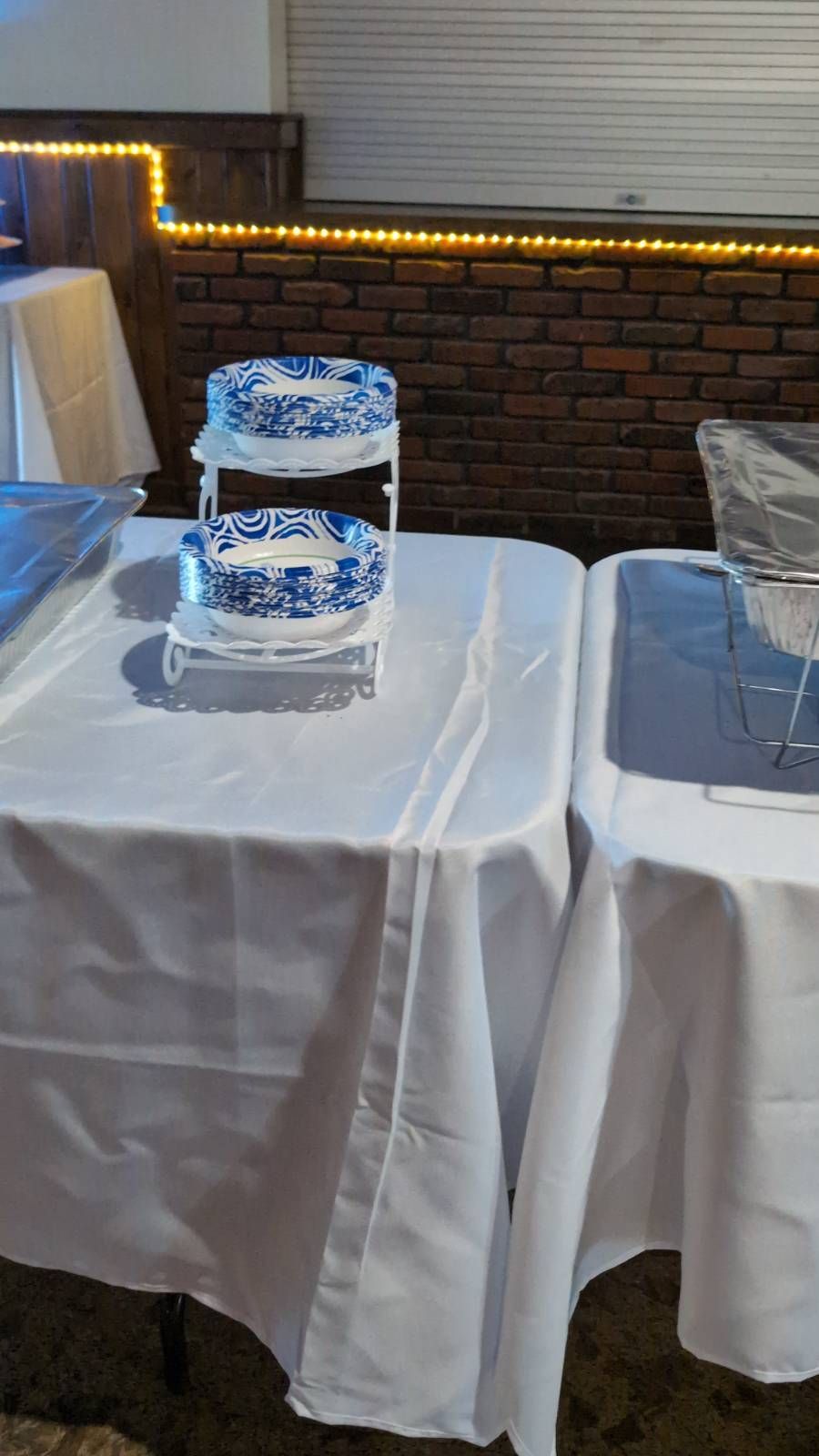 Glass water pitcher on a white tablecloth at an indoor event, with brick wall and warm string lights behind it