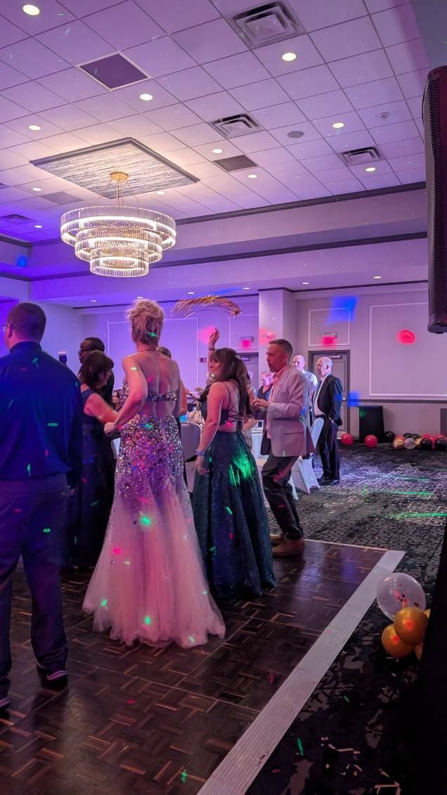 Guests in formal wear dance under purple lights in a ballroom with a chandelier.