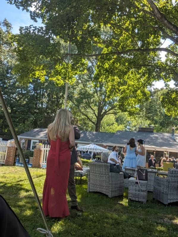 Outdoor gathering on a lawn with wicker seating, a tented table, and a woman in a red dress standing nearby
