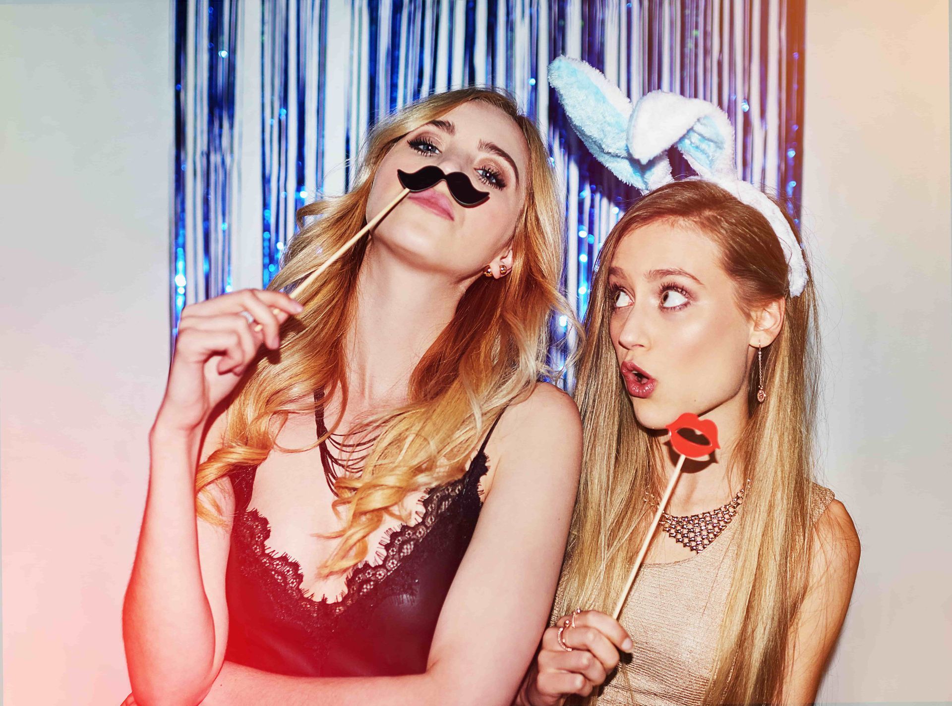 Two people pose with photo booth props—a mustache and red lips—in front of a blue tinsel curtain.