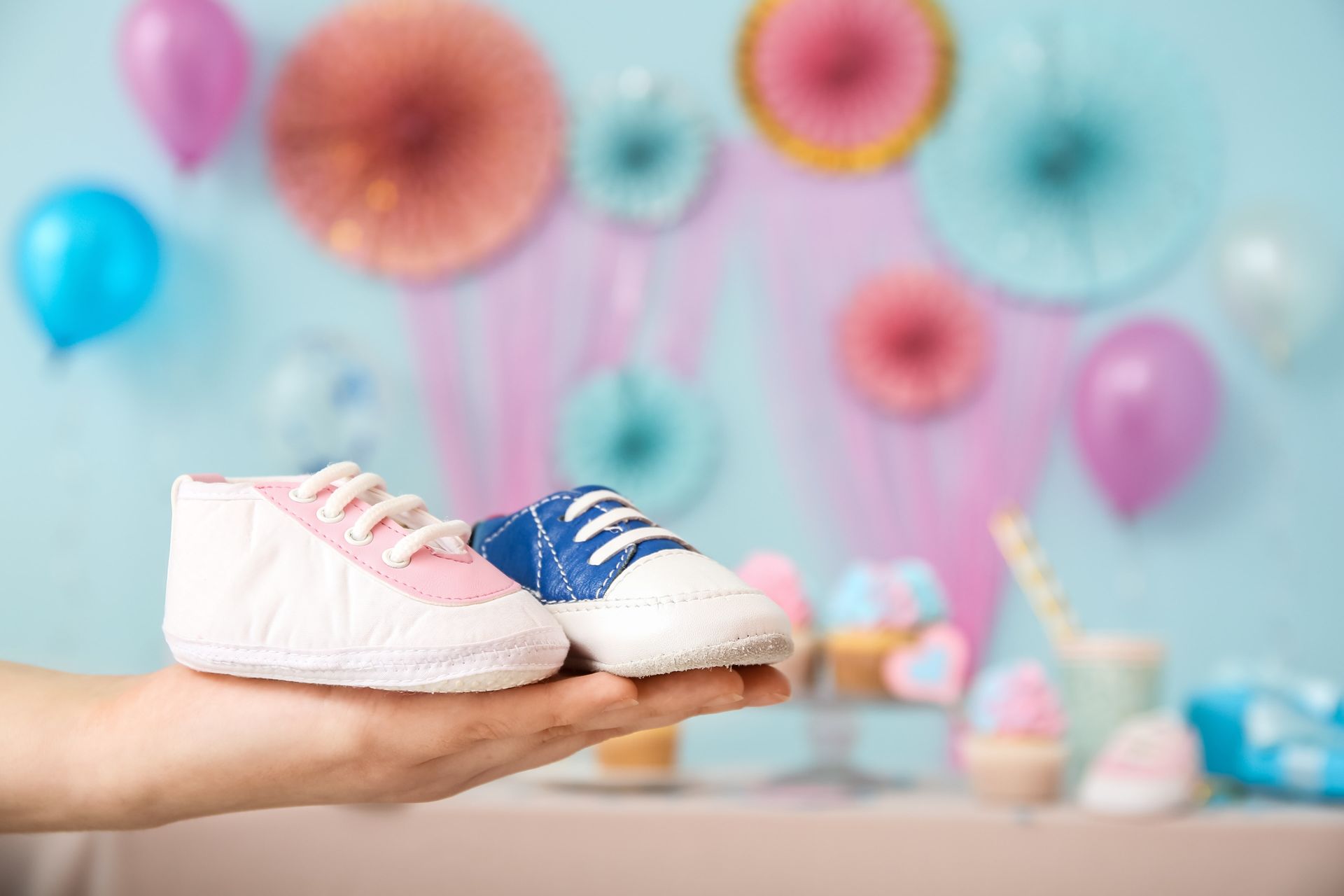 Hand holding pink and blue baby shoes in front of a baby shower backdrop with balloons and decorations.