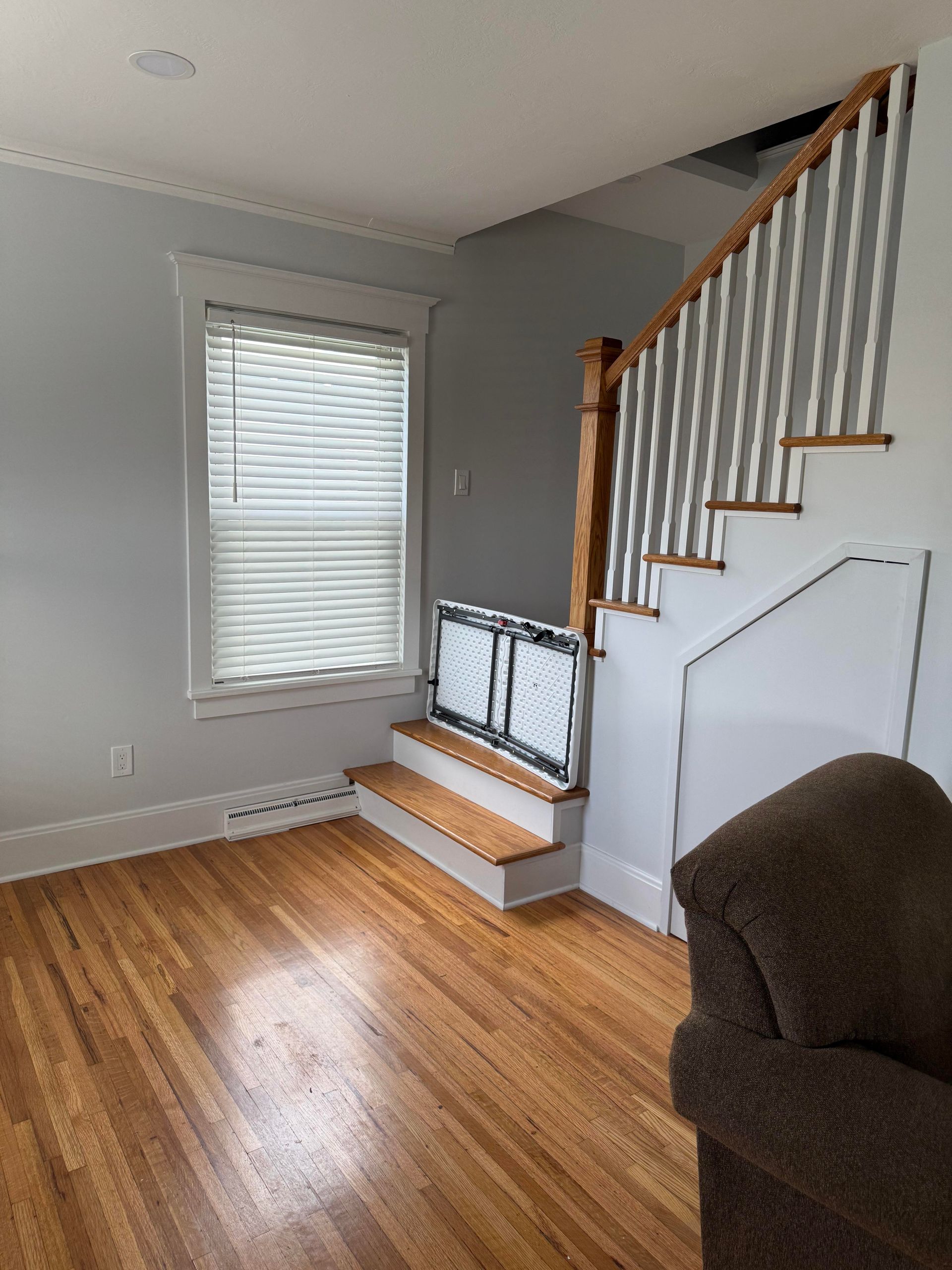 Empty living room with hardwood floors, window blinds, and a staircase with white railing.