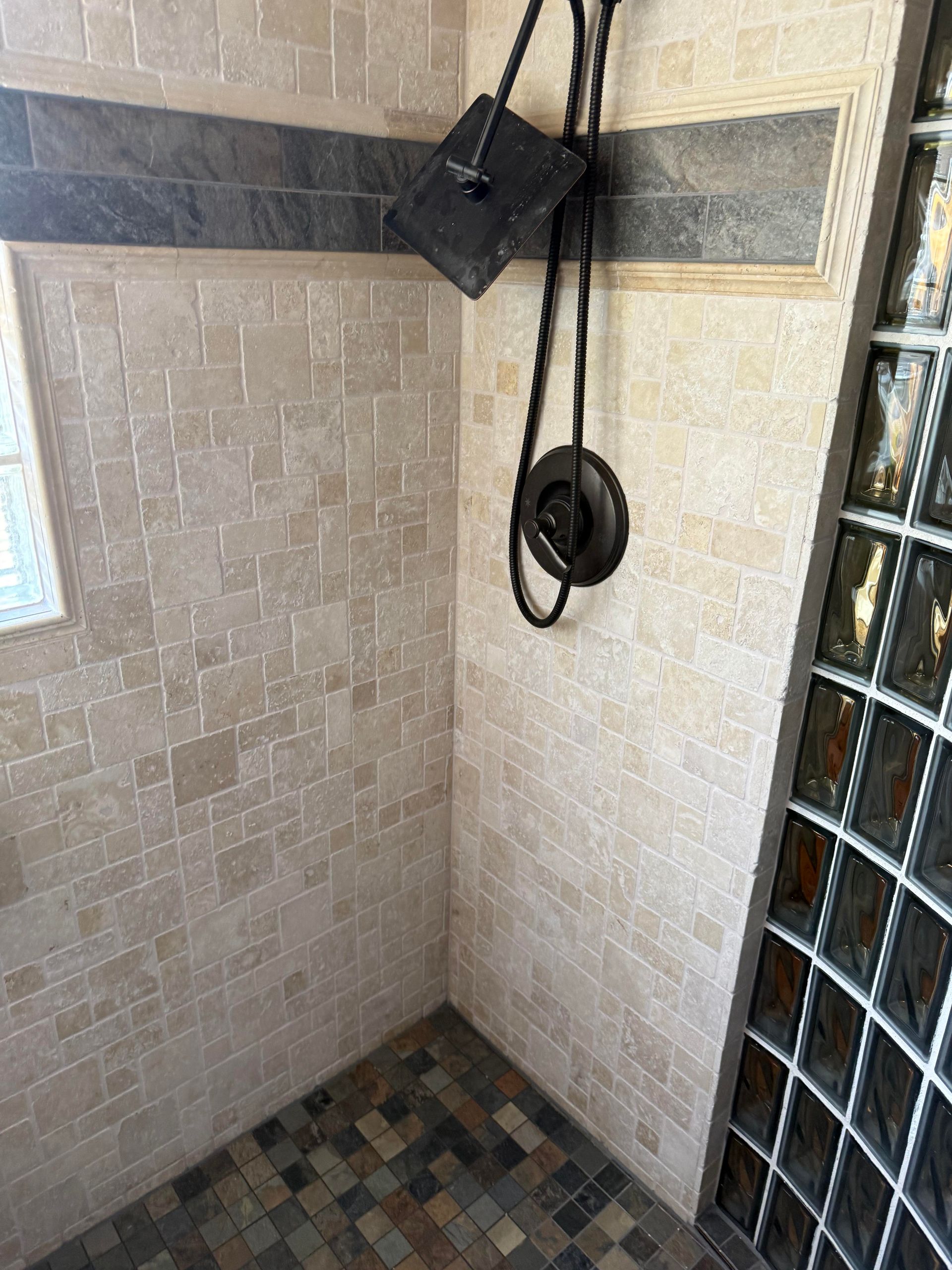 Small tiled shower corner with a hanging shower hose and dark trim near a glass block window.