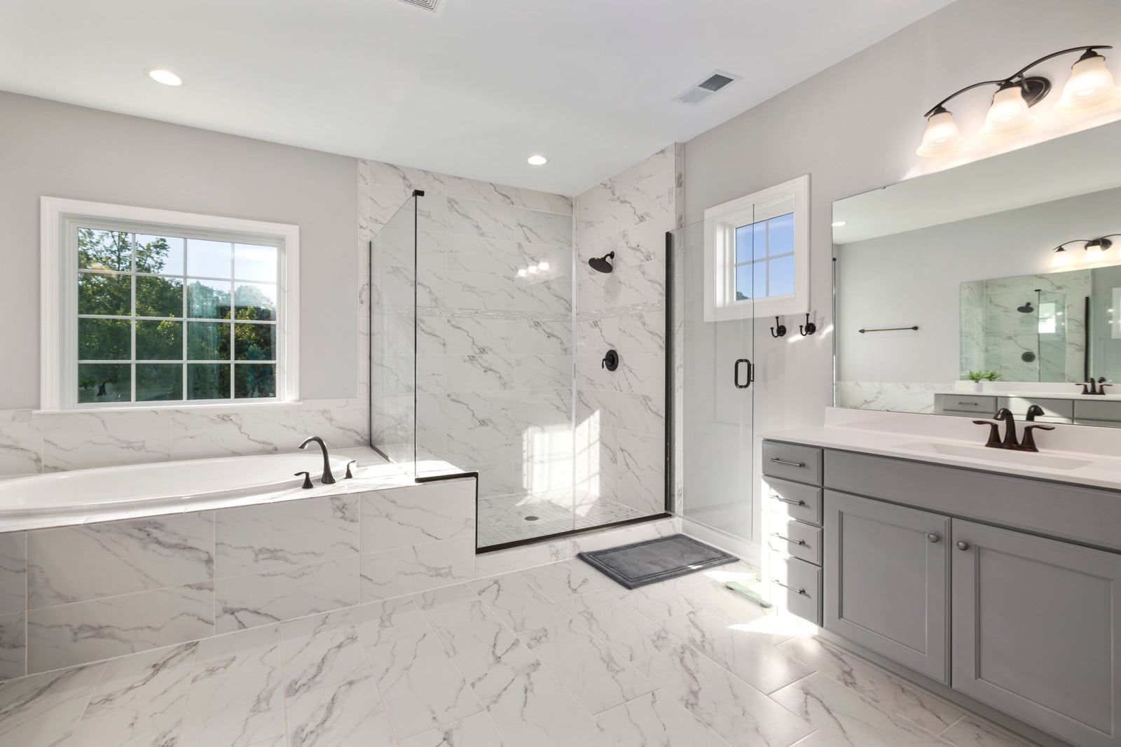 Bright modern marble bathroom with soaking tub, walk-in shower, gray vanity, and large mirror.