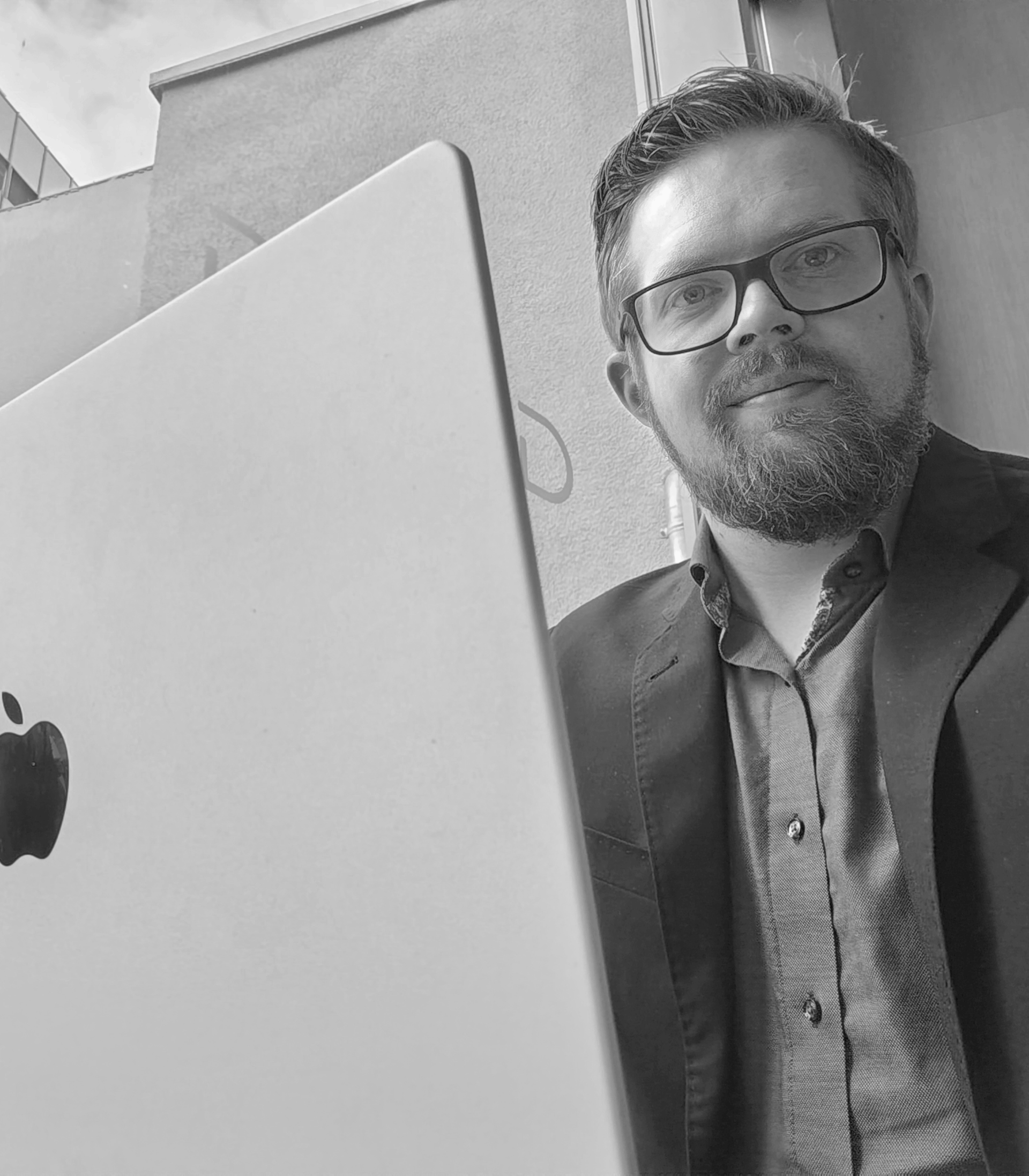 Mies seisoo suuren tietokoneen näytön takana silmälasit ja bleiseri päässä mustavalkoisessa valokuvassa