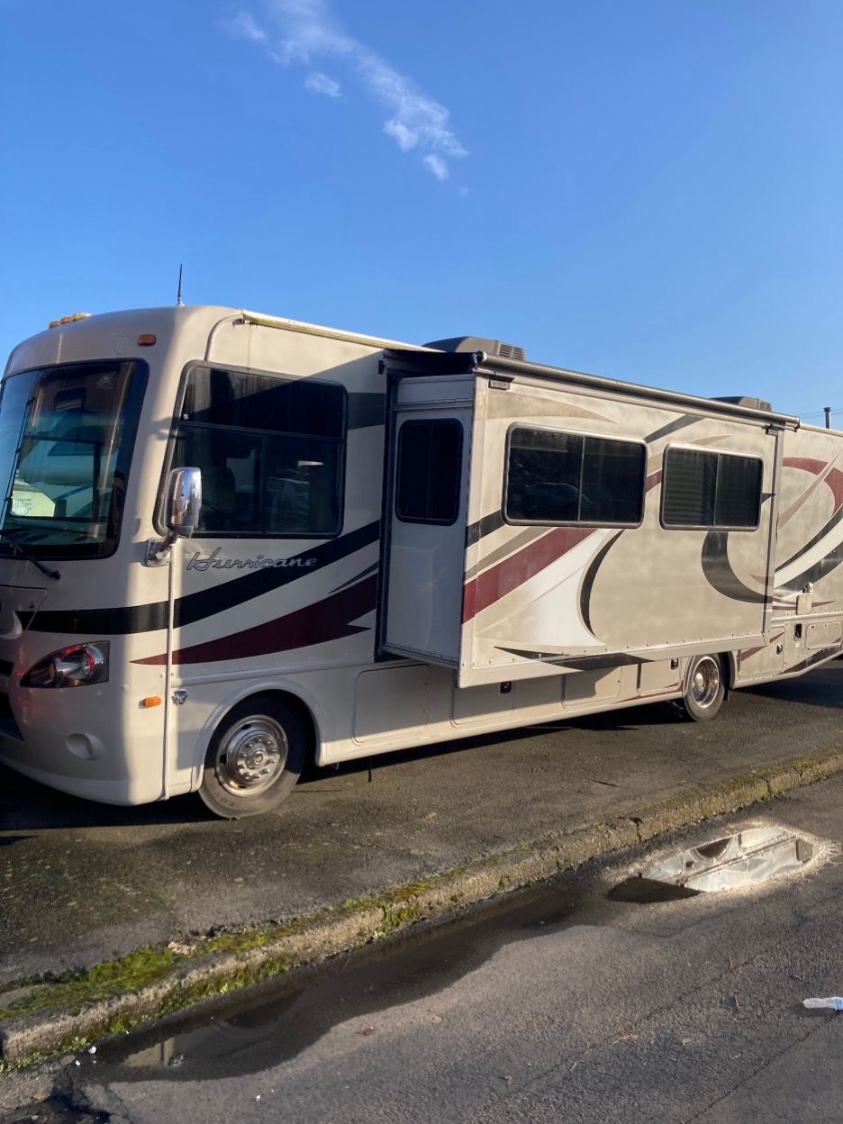 Tan and maroon RV parked on pavement, with slide-out extended, under a partly cloudy sky.