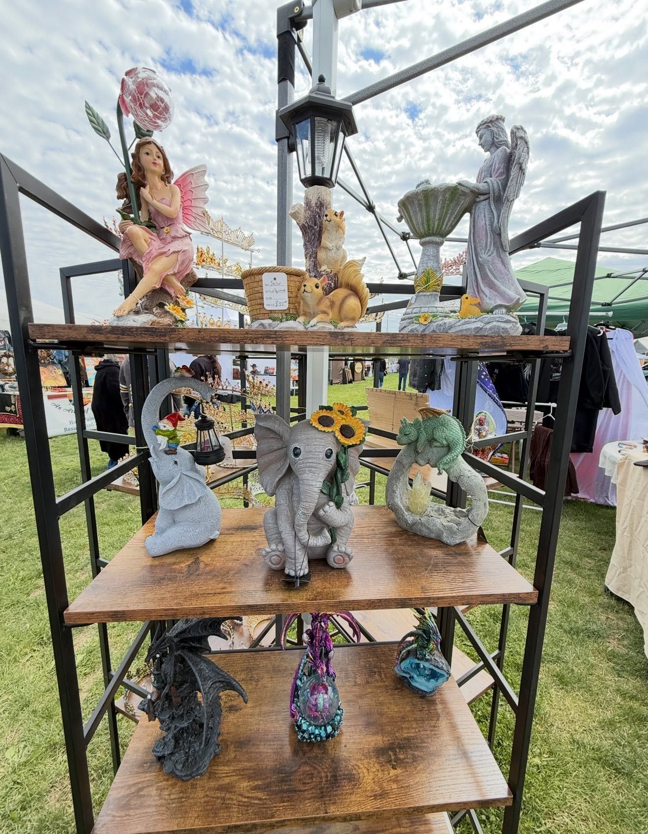 Outdoor display of colorful handmade figurines on tiered wooden shelves at a craft fair.