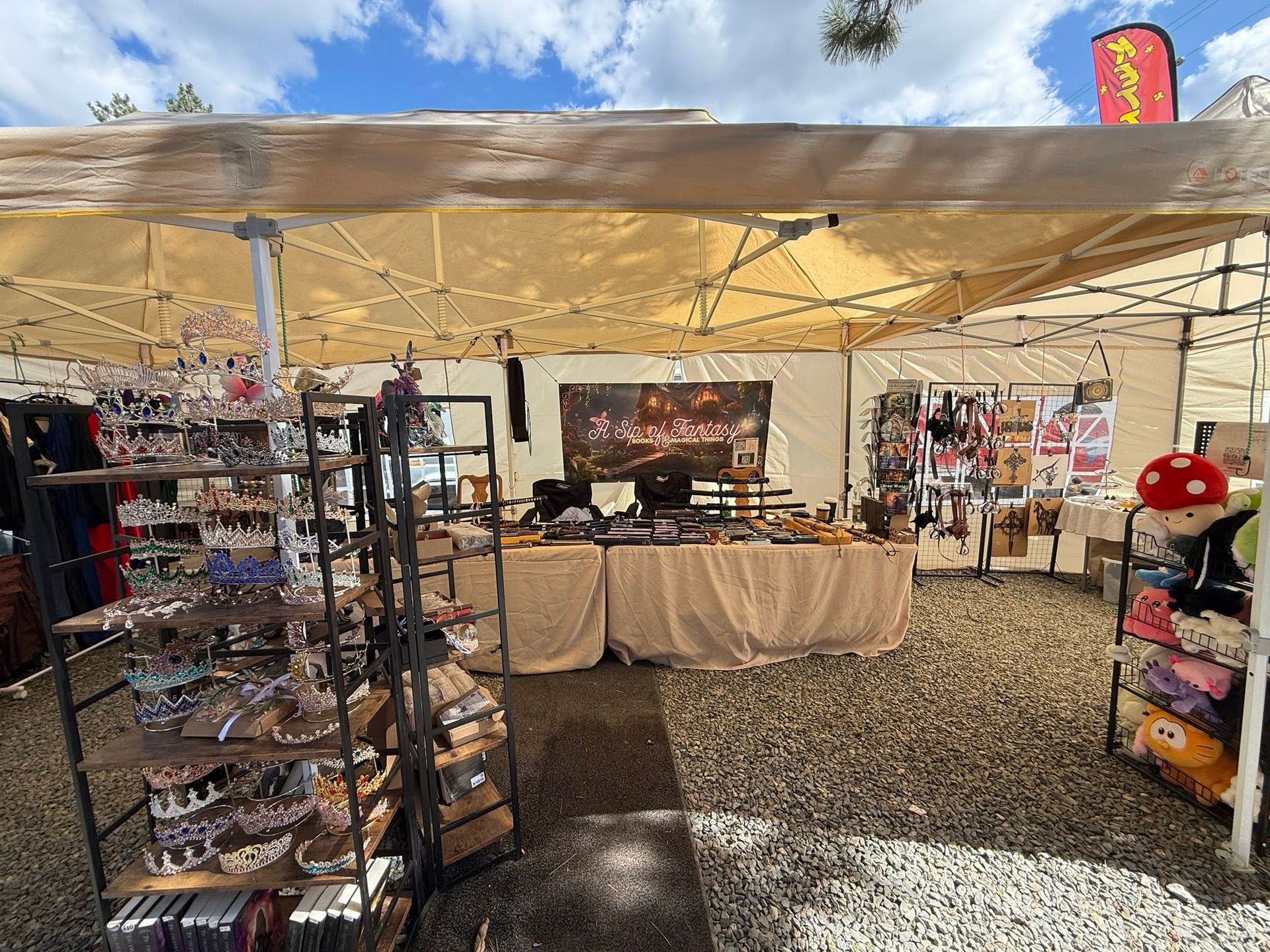 Outdoor market stall under a tan canopy, displaying jewelry and handmade crafts on metal racks and tables.
