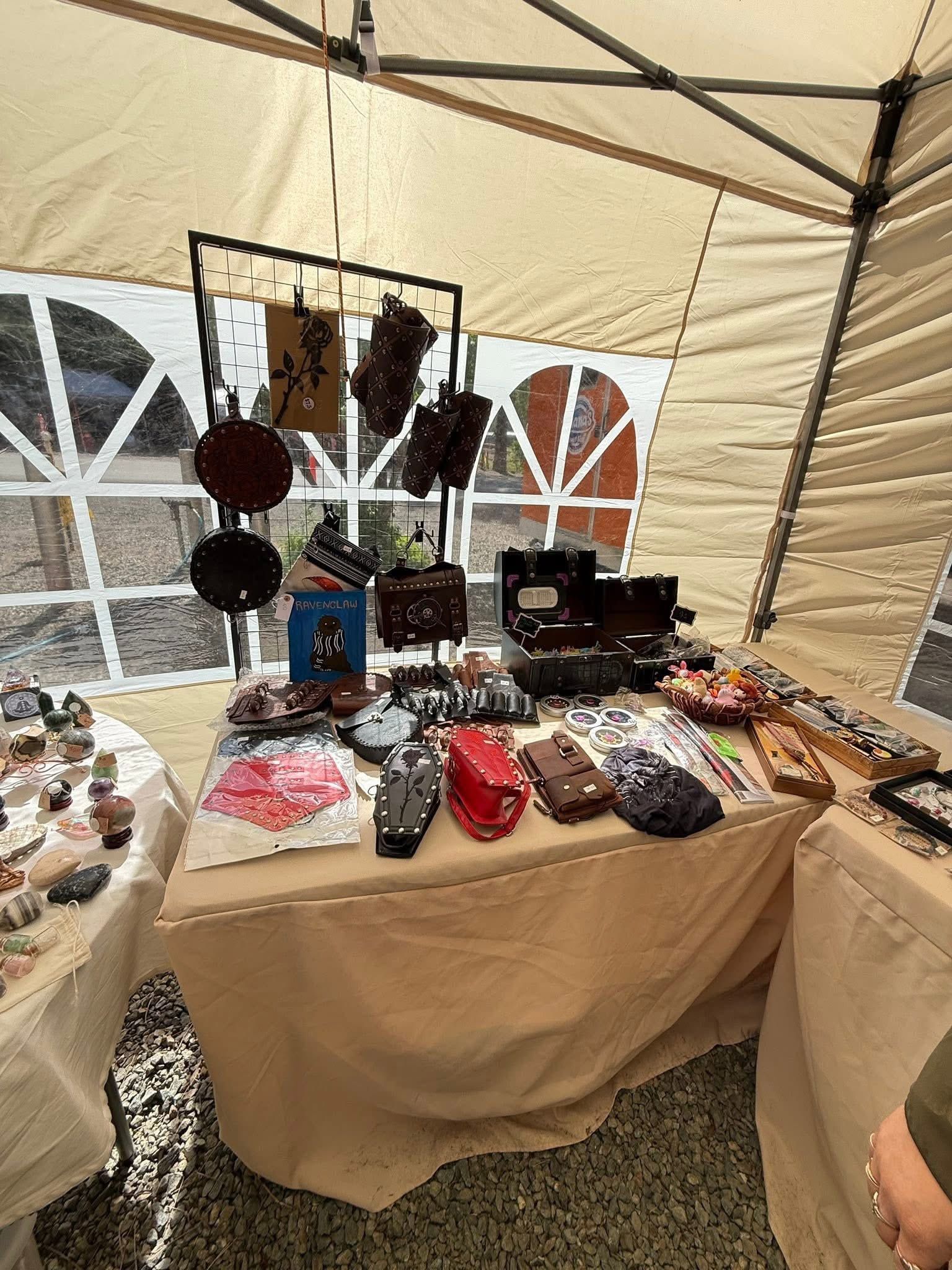 Craft fair table under a beige tent, displaying handmade jewelry, accessories, and small packaged goods.