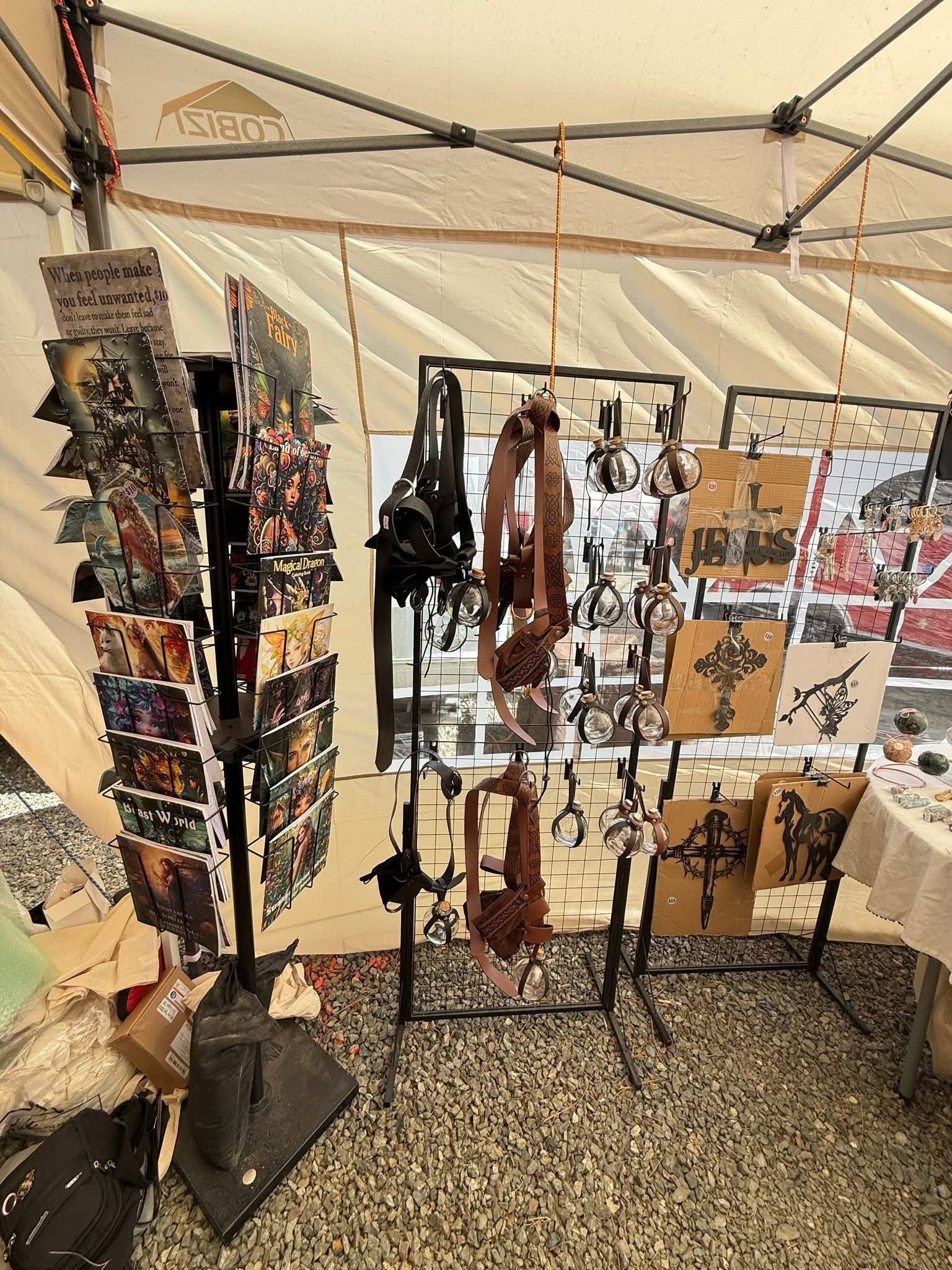 Outdoor market display of handmade jewelry, photos, and crafts on stands under a white tent