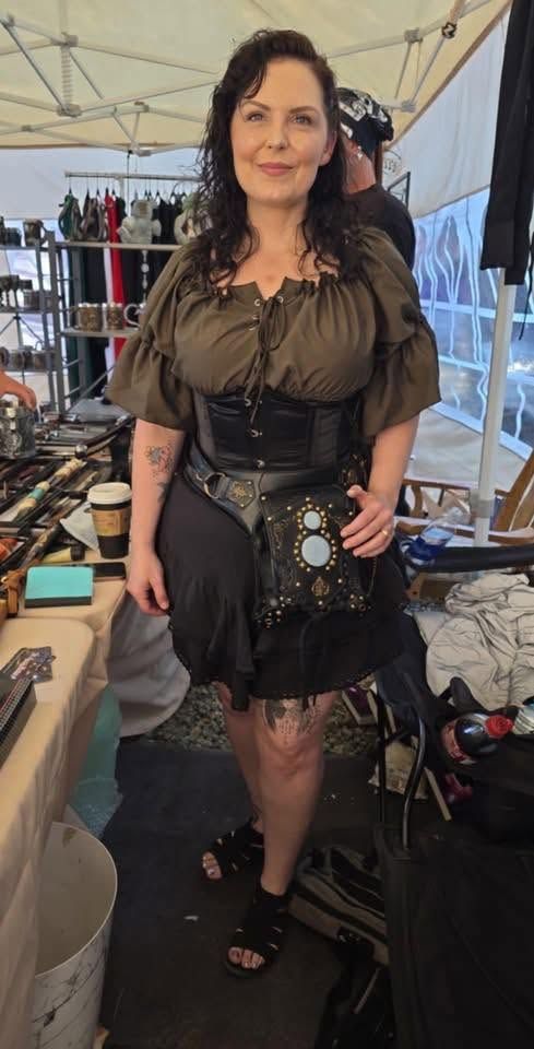 Woman in dark dress and boots standing at an outdoor market stall with handmade items displayed.