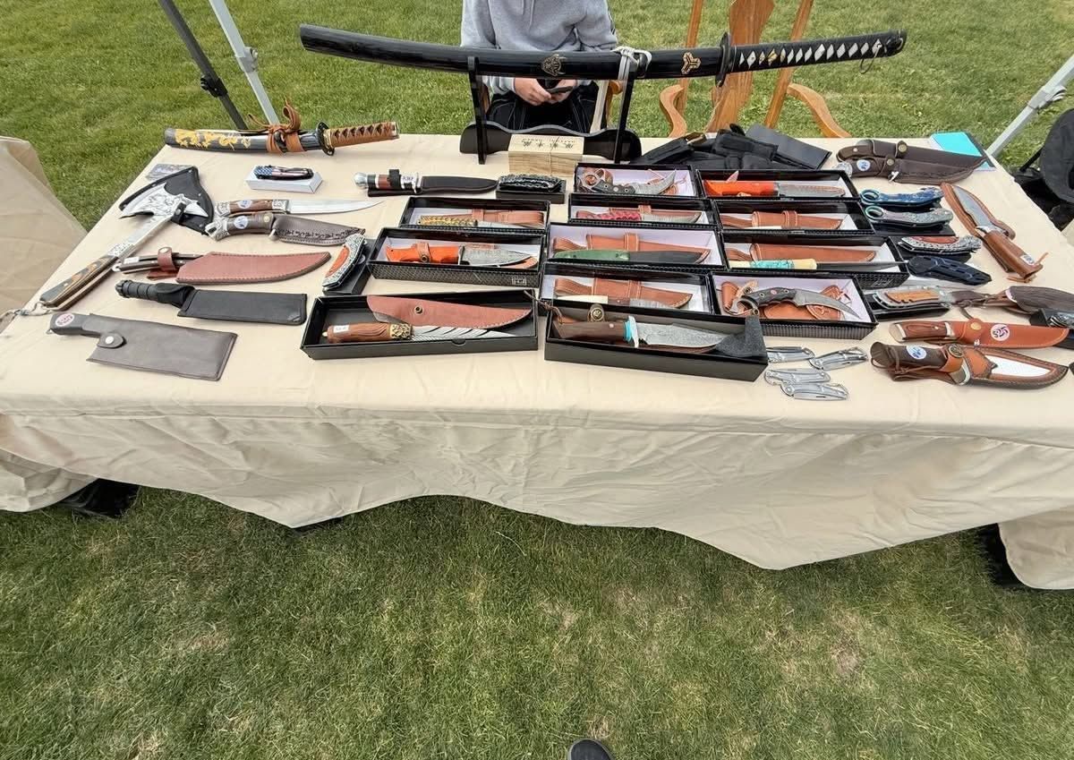 Table covered with rifles, pistols, and gun cases displayed outdoors on grass.