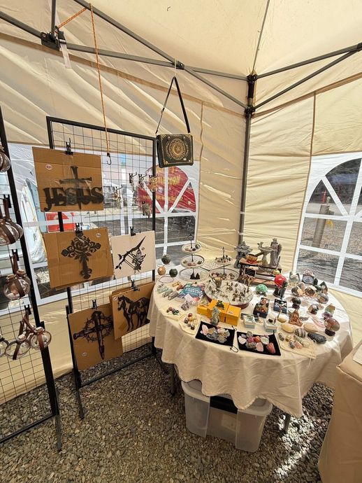 Small vendor booth with a round display table of crafts and hanging photo prints inside a beige tent.