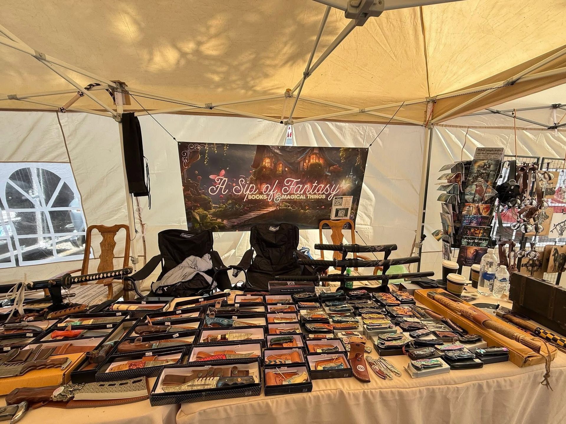 Vendor stall under beige tent displaying rows of knives and tools on tables, with a dark banner behind.