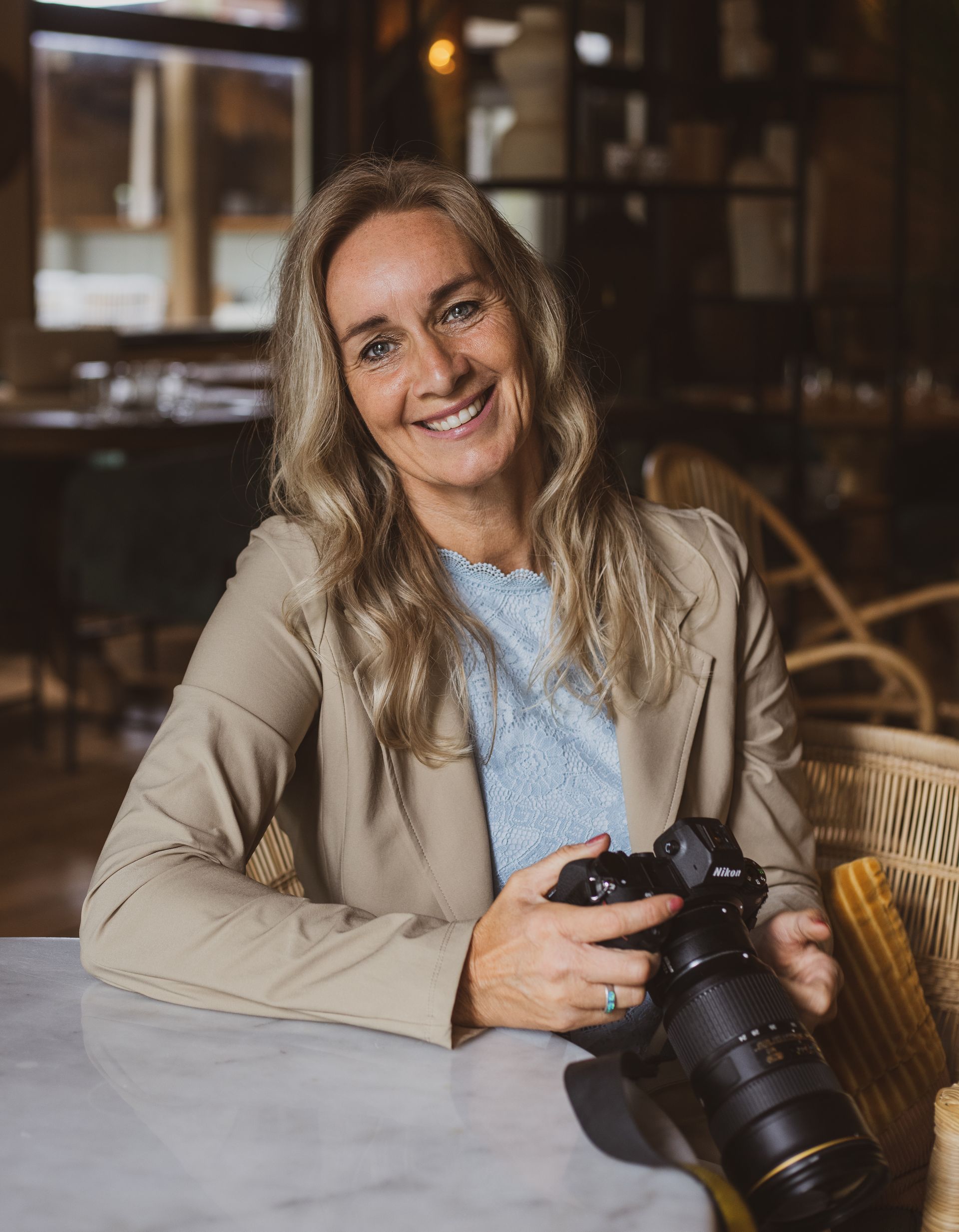 Een vrouw zit aan een tafel, houdt een camera vast en glimlacht.