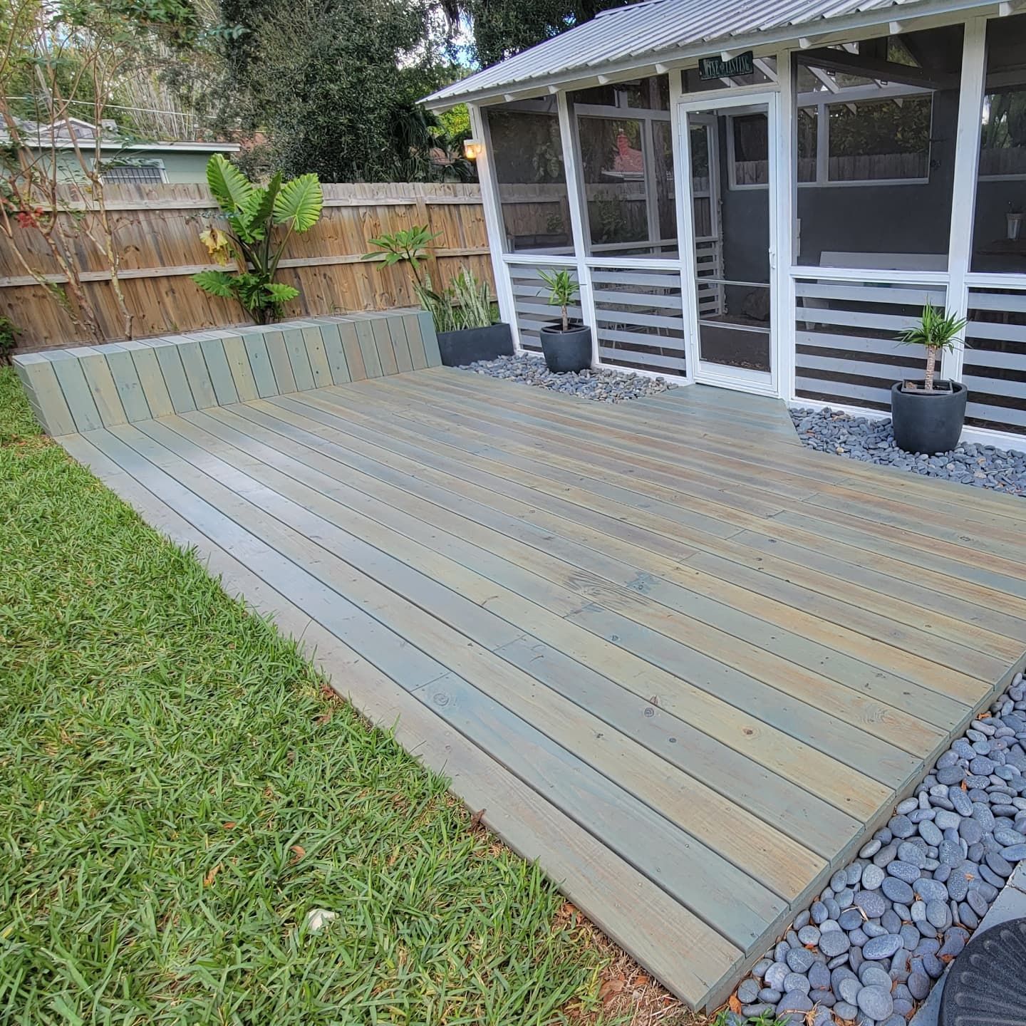 Backyard deck with gray boards beside a screened patio, bordered by grass and gravel.