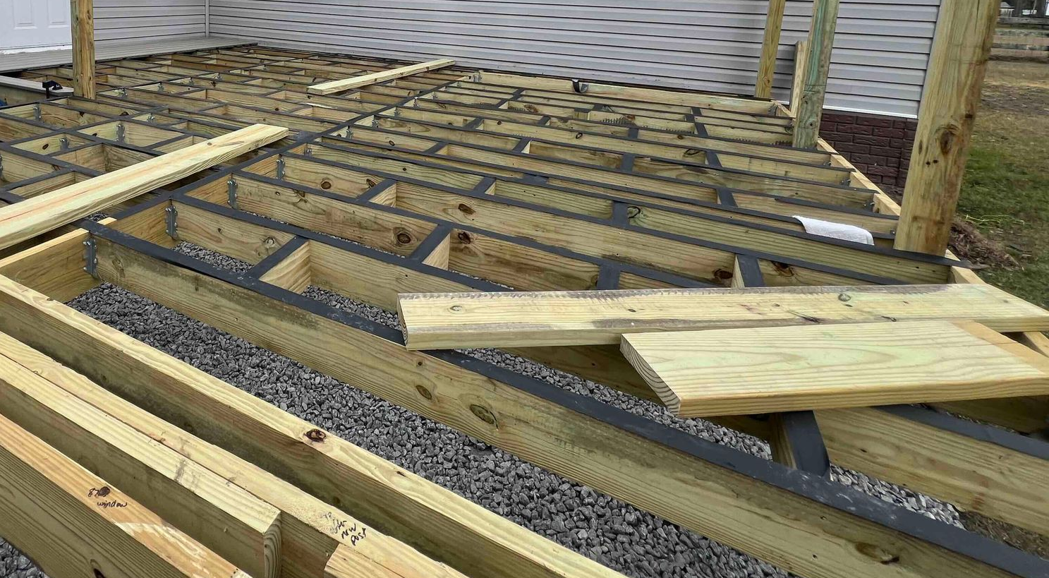 A wooden deck frame under construction with gravel underneath, near the exterior wall of a house.