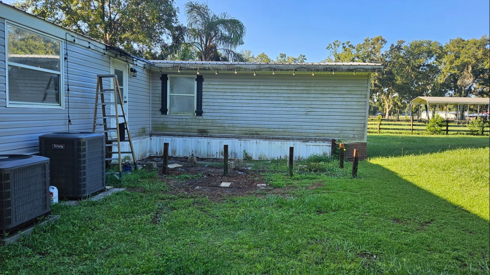 A single-story mobile home with light blue siding, a metallic side extension, two AC units, and a ladder by the doorway.