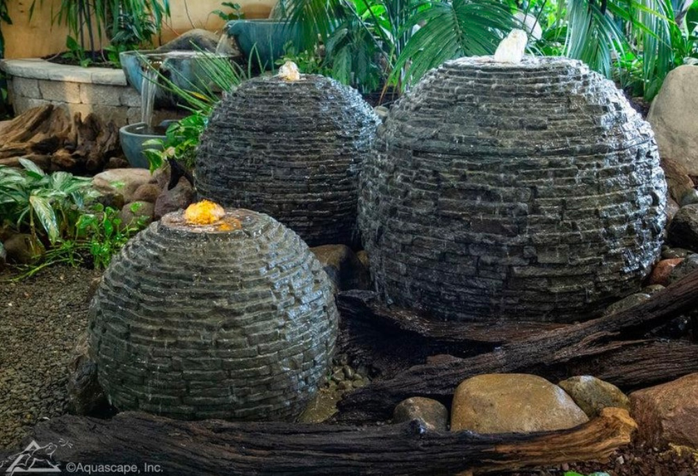 Three stone sphere fountains with water spouts, set among rocks and plants.