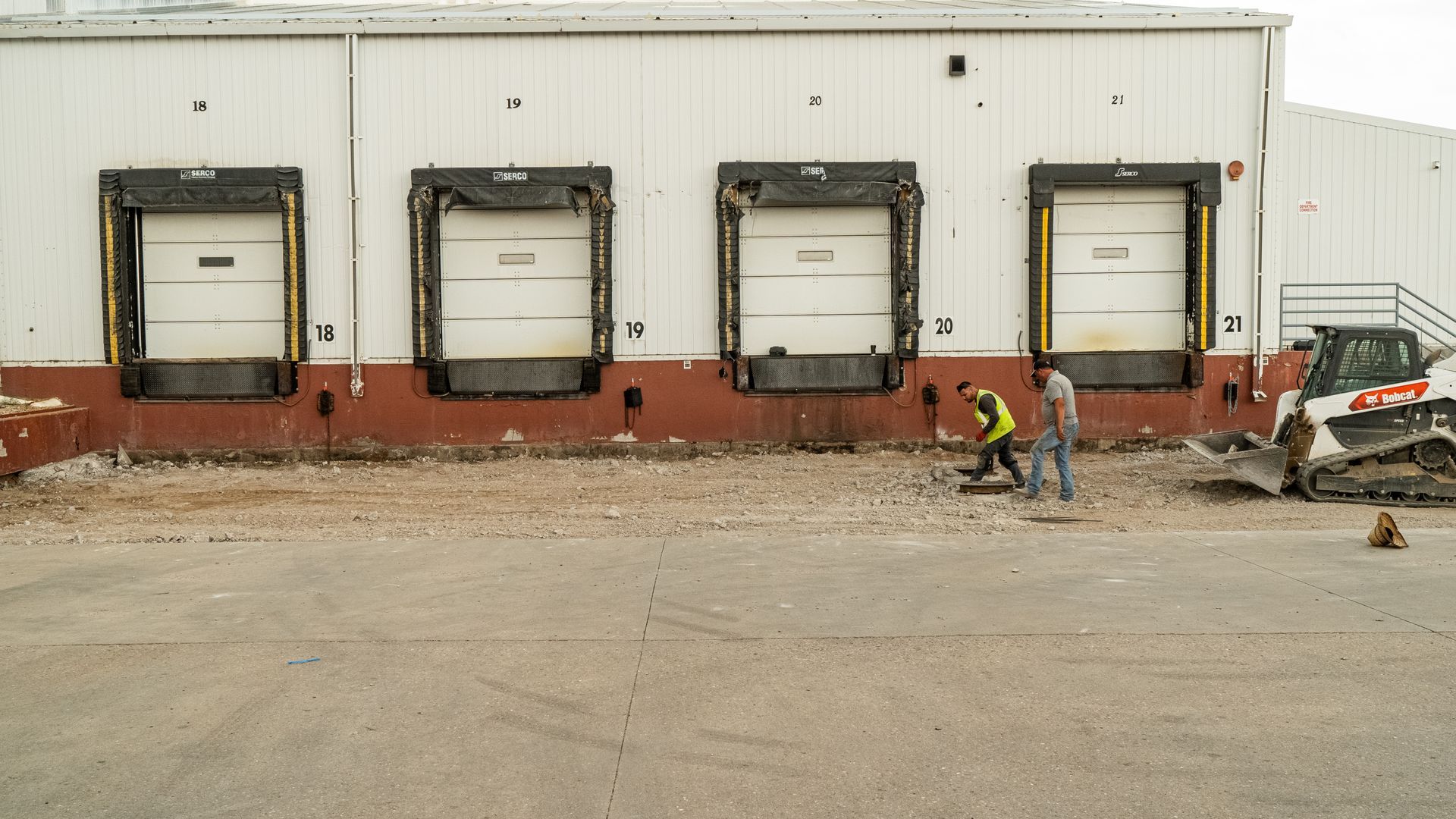 Workers stand near loading docks on an industrial building beside a skid steer and debris.