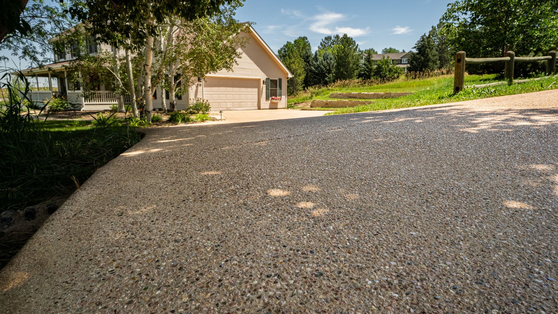 Gravel driveway leading to a beige house with trees and green lawn on a sunny day