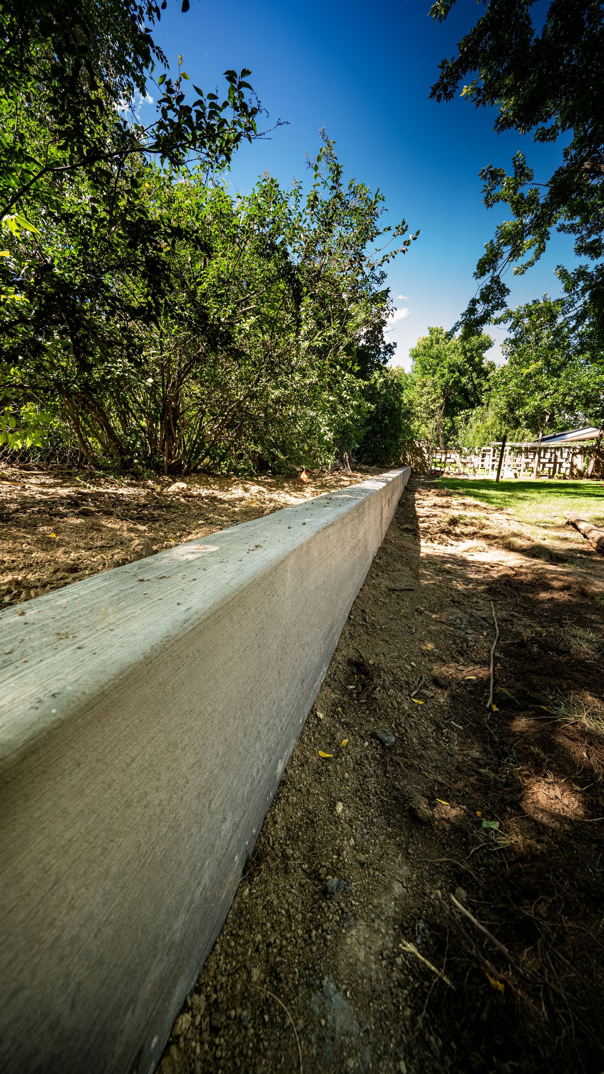 Long white concrete wall beside a leafy orchard under a blue sky