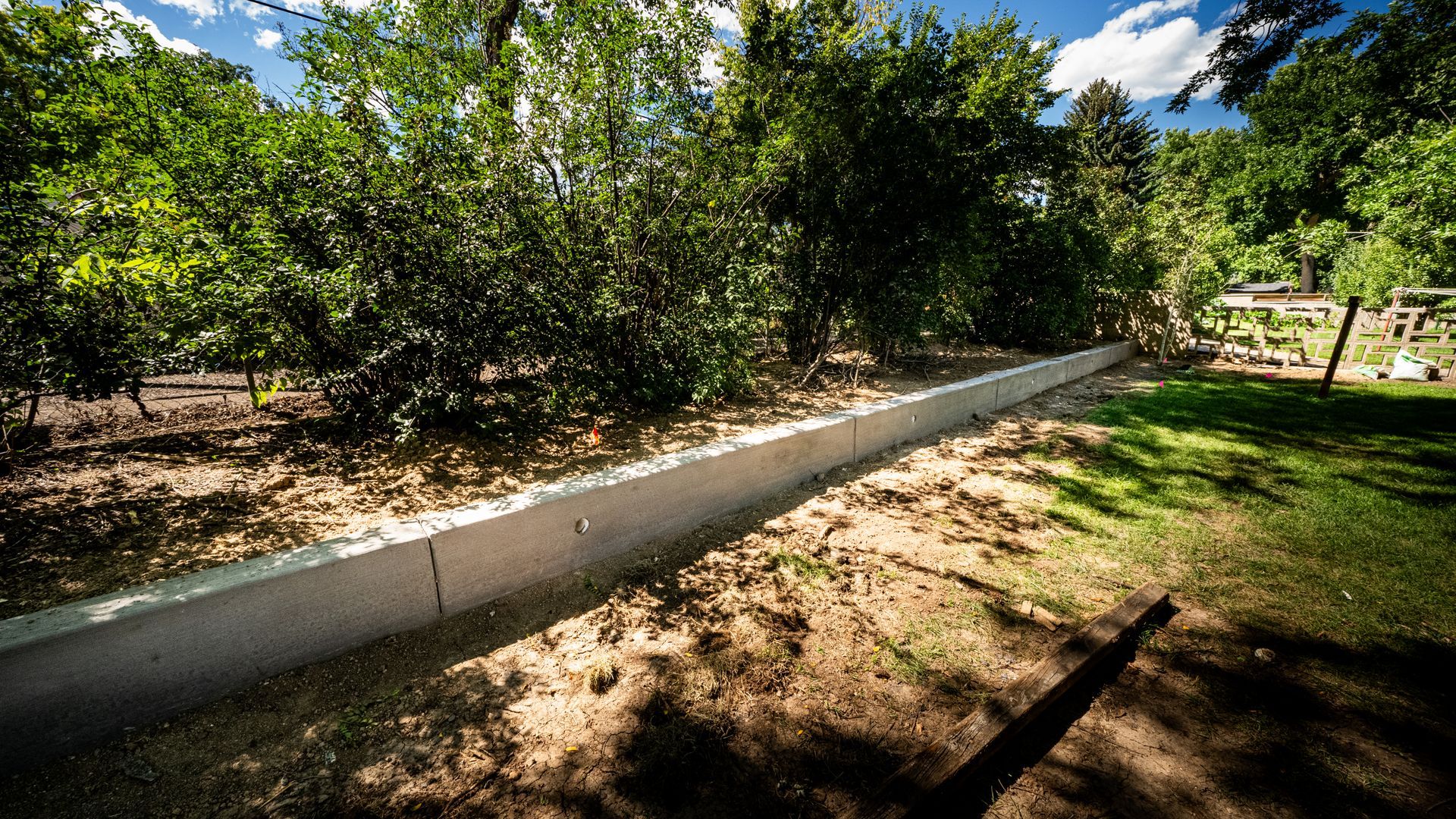 Concrete retaining wall along a dirt path beside leafy trees and a bright blue sky.