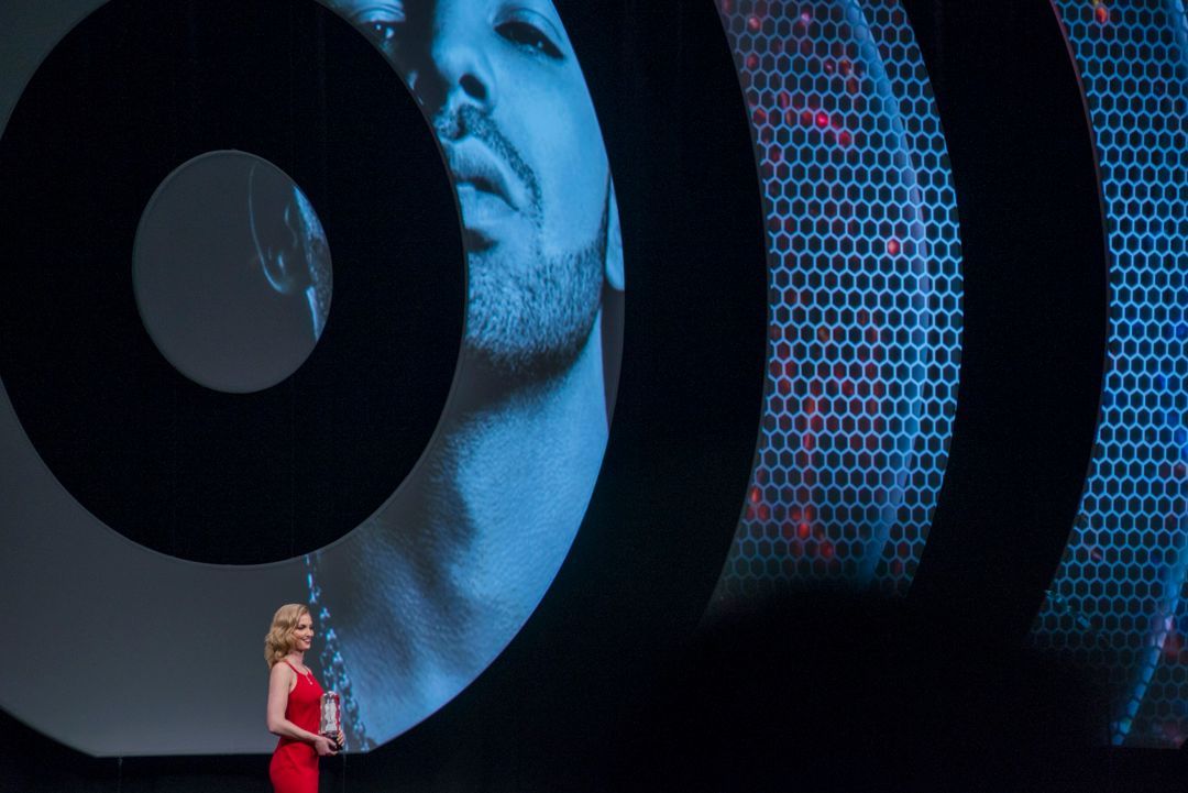 A woman in a red dress is standing in front of a large screen with a picture of a man on it.