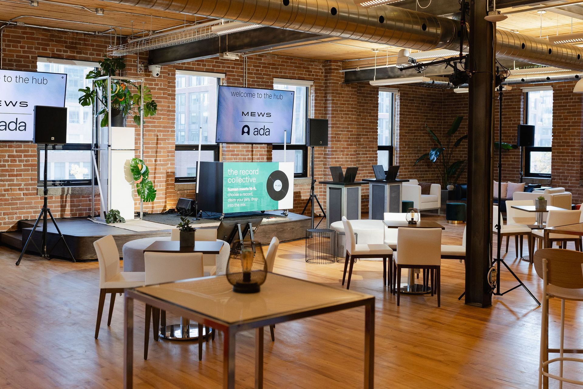 Event space with tables, chairs, and stage against exposed brick walls. Several screens display text.