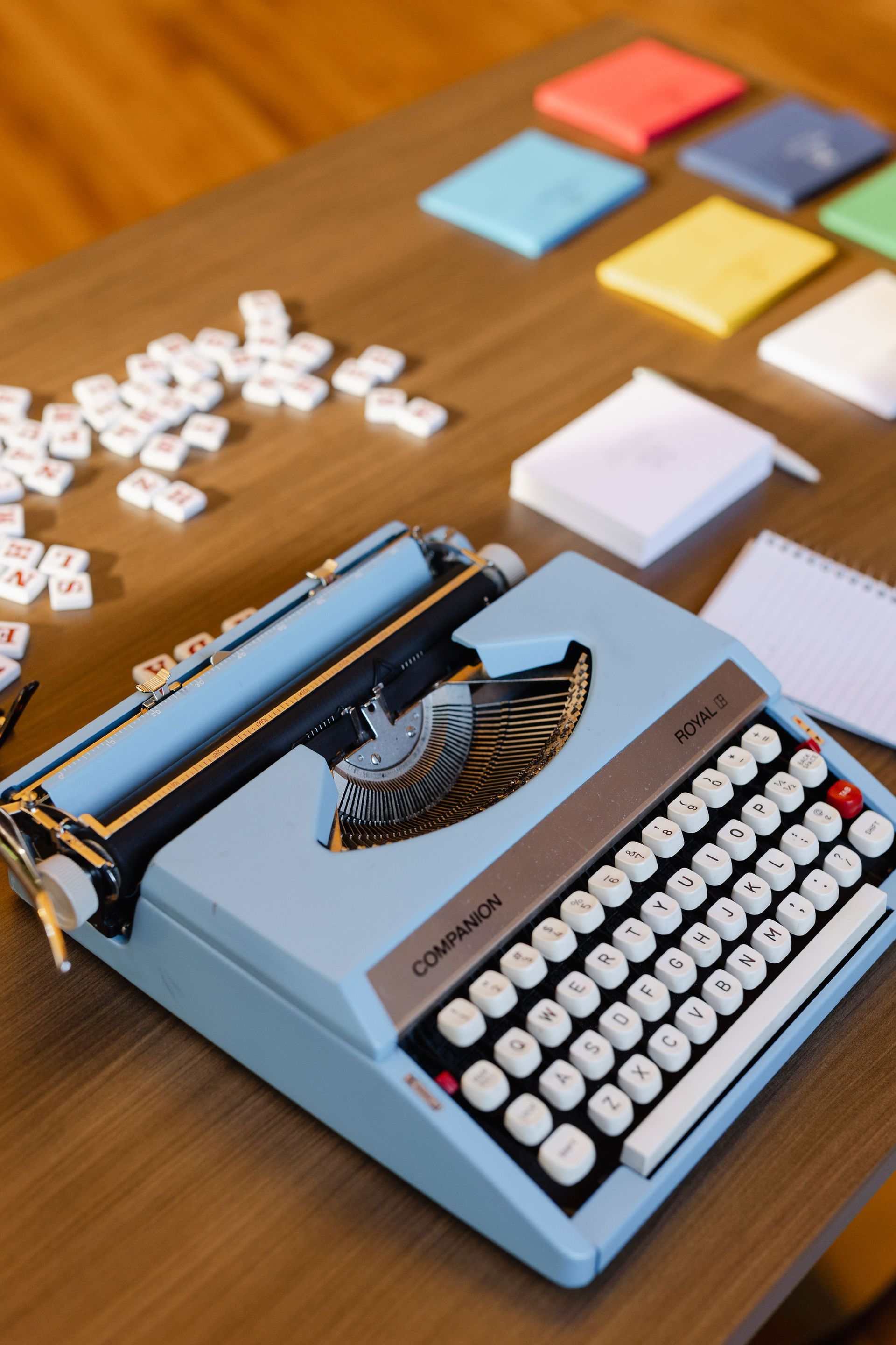 Blue typewriter with white keys on a wooden table, scattered tiles, small notepads, and colorful squares in the background.
