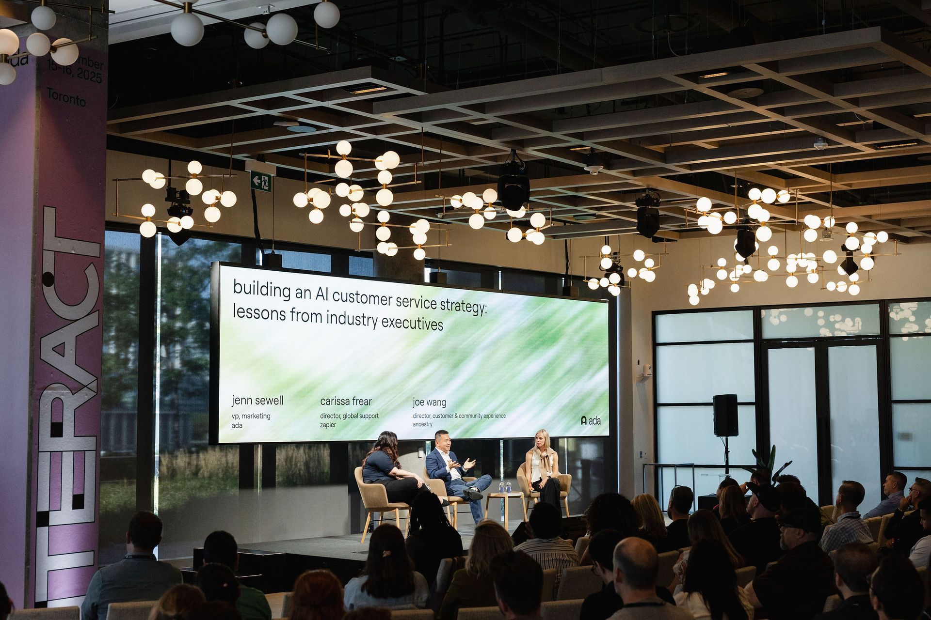 Panel discussion on stage with audience, large screen, bright overhead lights, and a glass-walled venue.