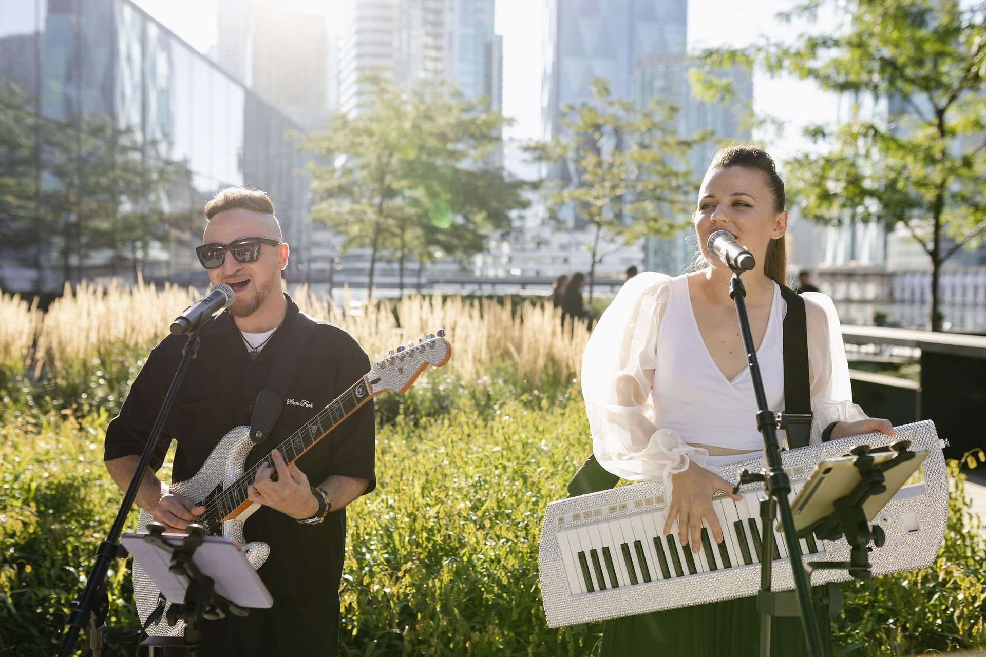 A duo performs outdoors: guitarist in sunglasses, and keyboardist singing, in front of a modern city backdrop.