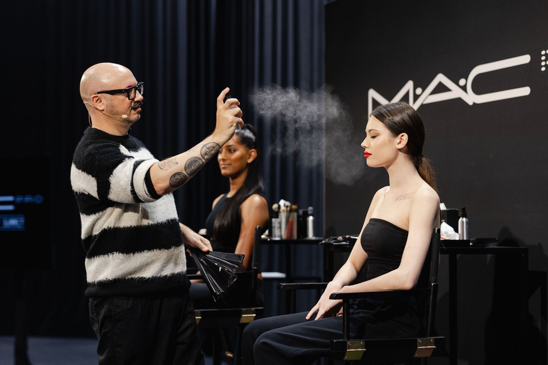 Makeup artist spraying a model's face at a MAC Cosmetics event; a second model watches in the background.