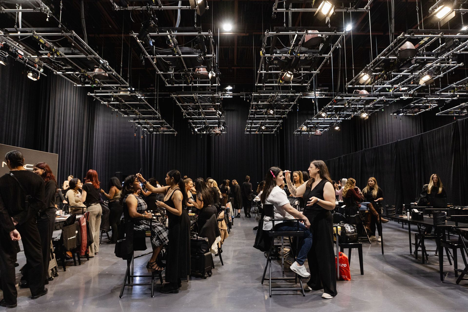 Makeup artist gestures toward model seated in a chair, both on a stage.