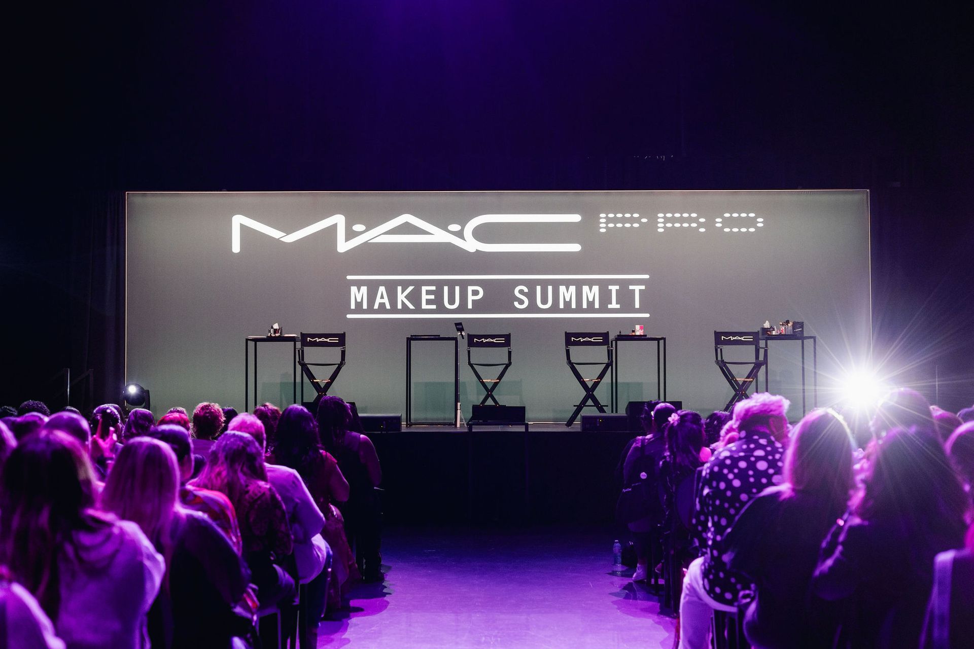 MAC Pro Makeup Summit stage with audience and logo. Purple lighting; stage setup with chairs.