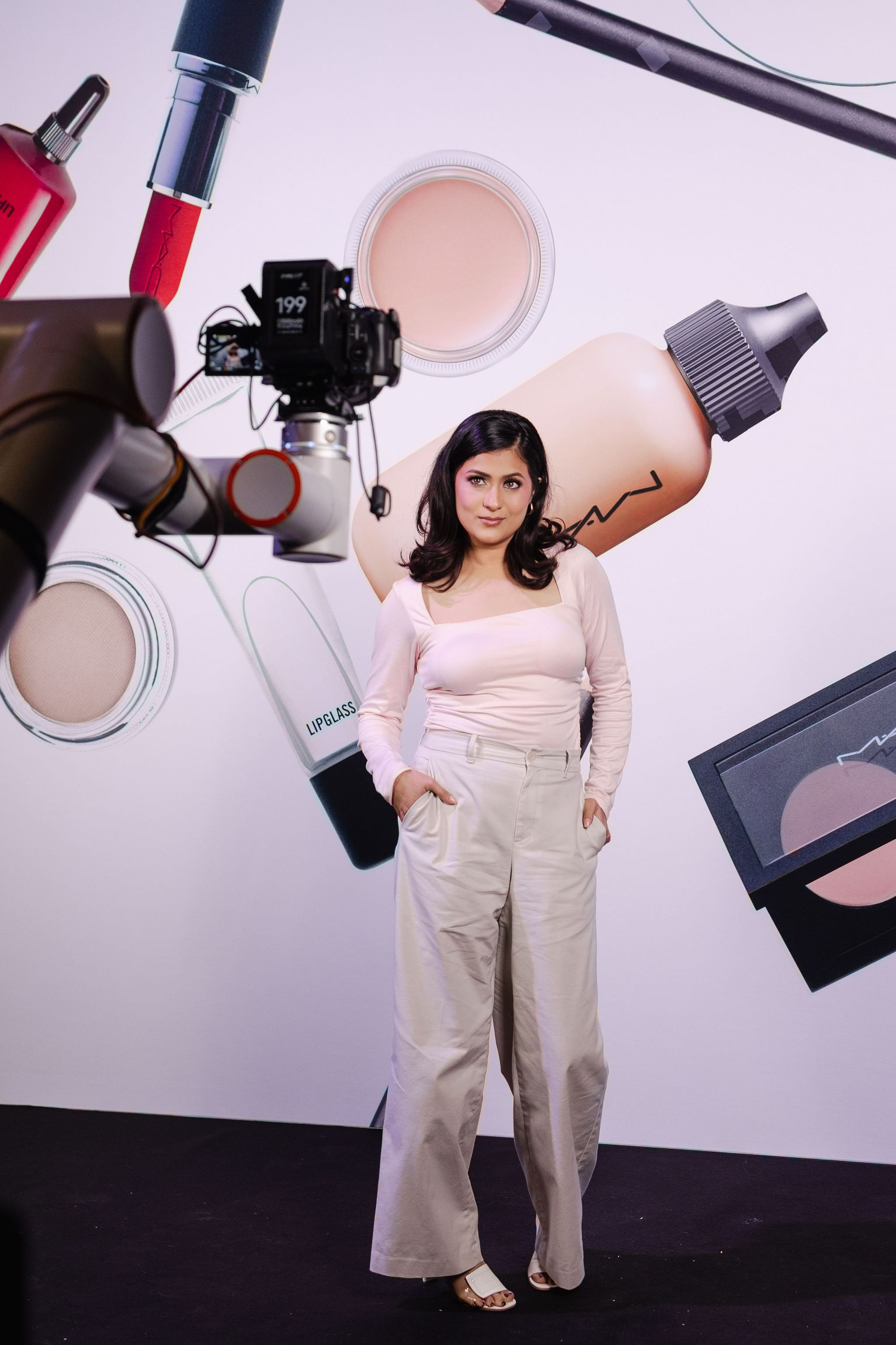 Woman in light-colored outfit standing in front of a makeup-themed backdrop. A camera is set up near her.