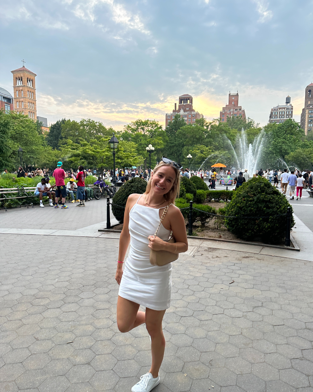 A woman in a white dress is standing in a park with a fountain in the background.