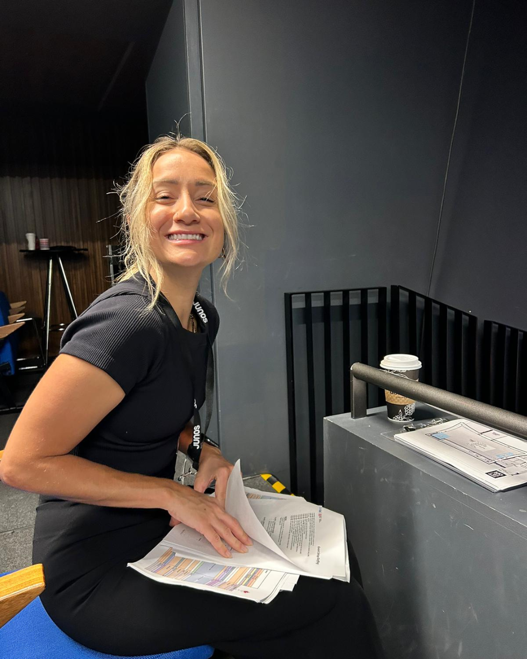 A woman is sitting on a blue chair holding papers and smiling.