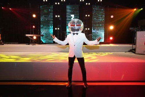 A man in a tuxedo is standing on a stage with a disco ball on his head.