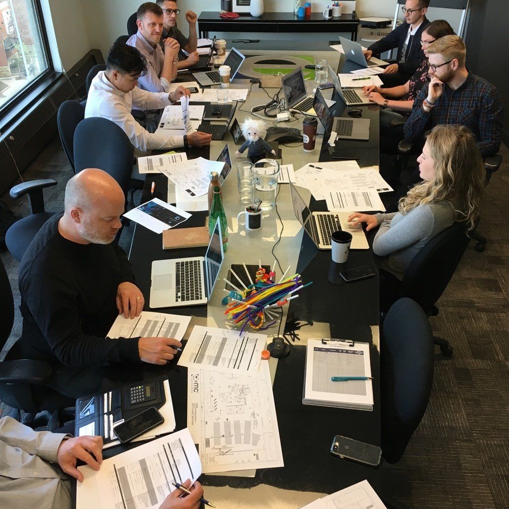 A group of people are sitting around a long table with laptops and papers