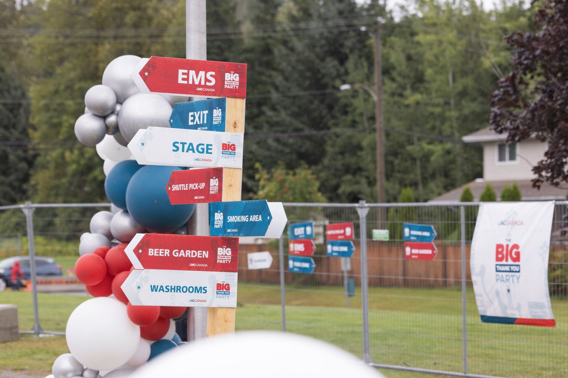 Decorated signpost with event labels and balloons at an outdoor fairground, with banners in the background