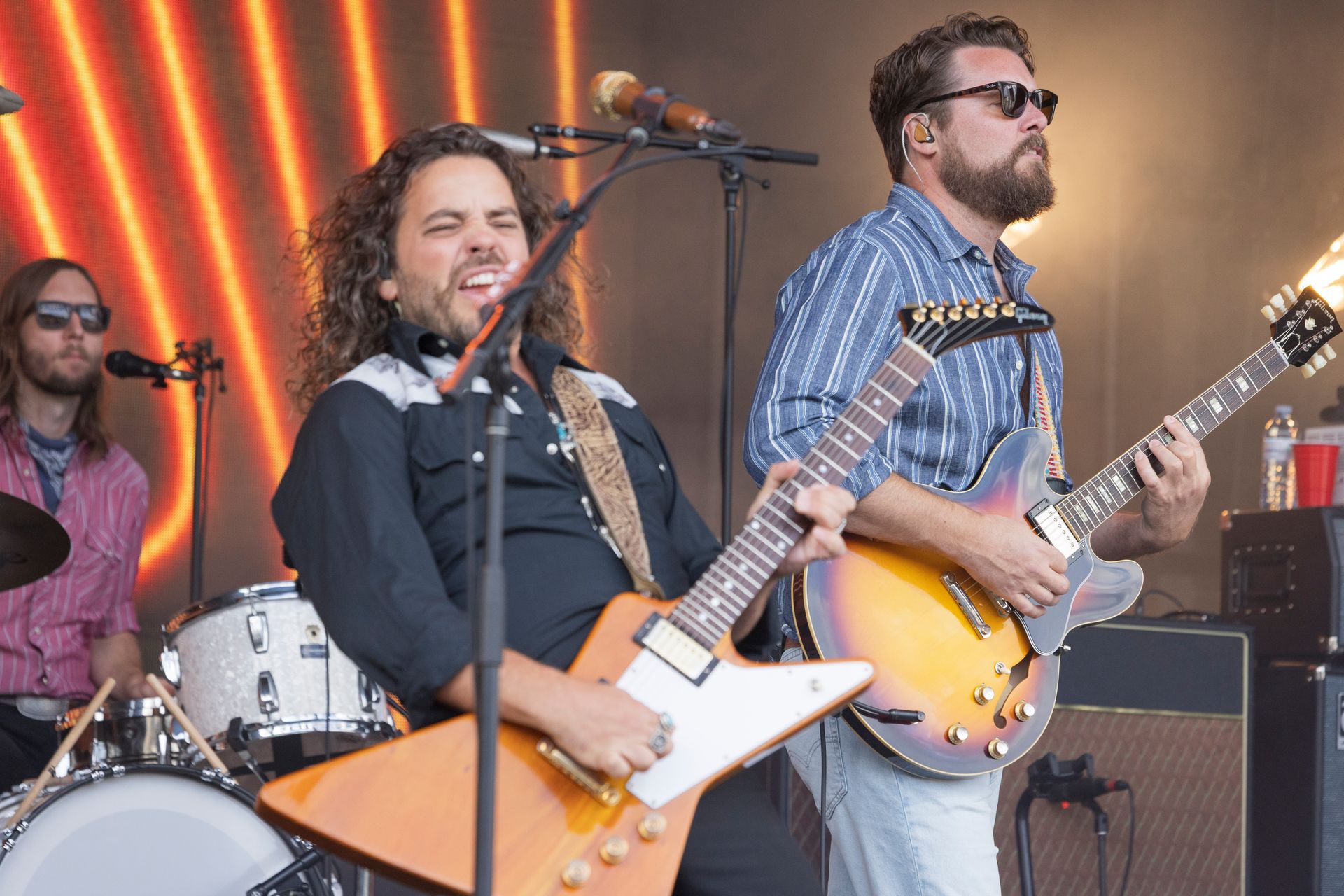 Band playing guitars on outdoor stage; the lead singer with long hair and a cowboy shirt is singing into a microphone.