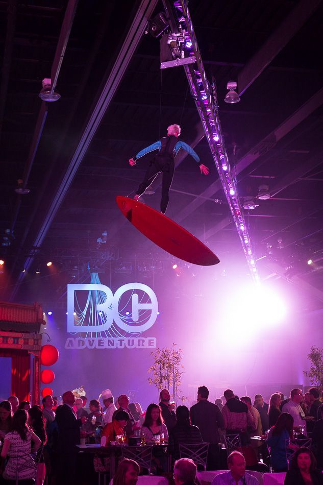 A person on a surfboard suspended in the air above a crowd at an event. Purple lighting, ABC Adventure logo.