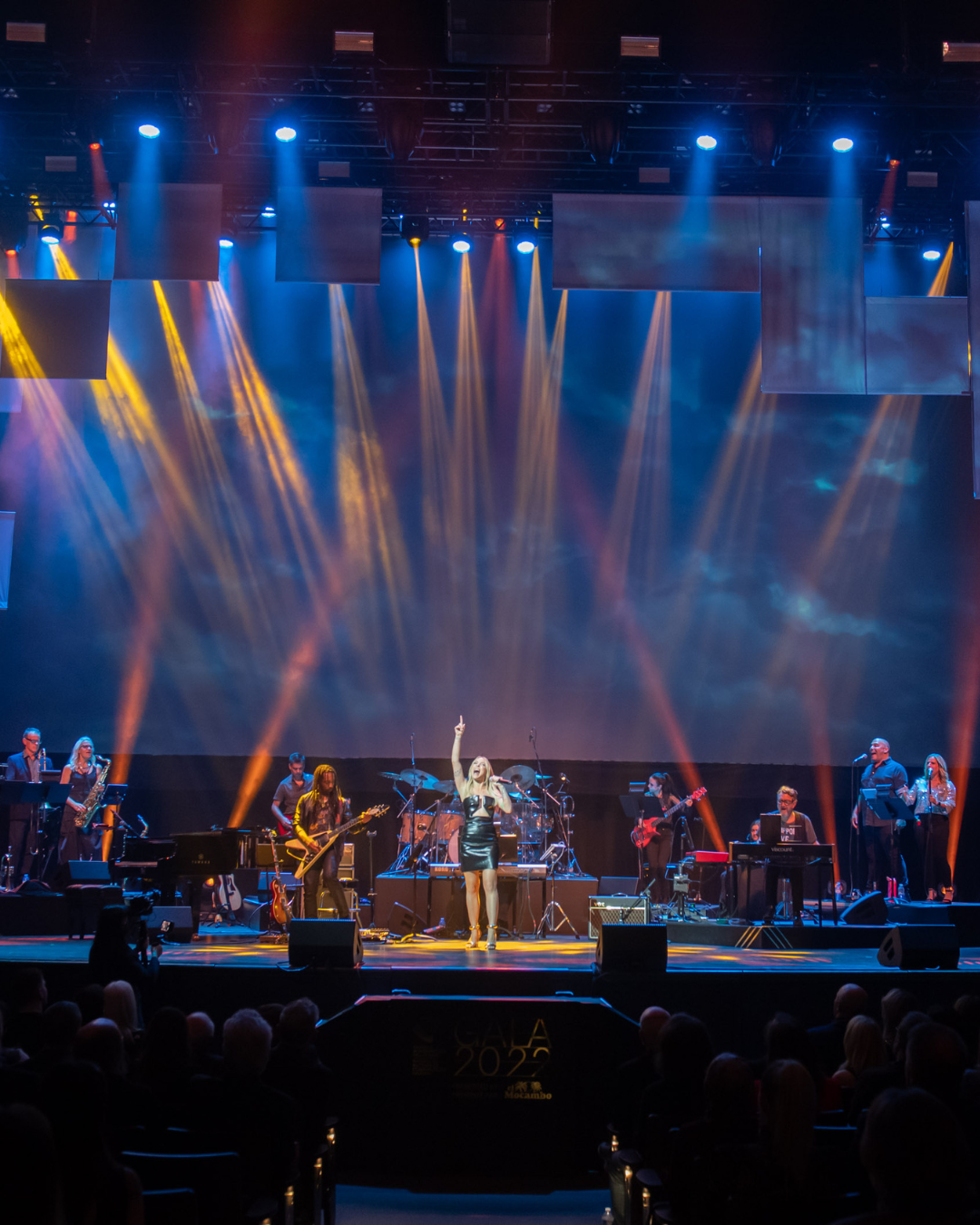 Live band performing on a stage with blue and orange spotlights and an audience in front