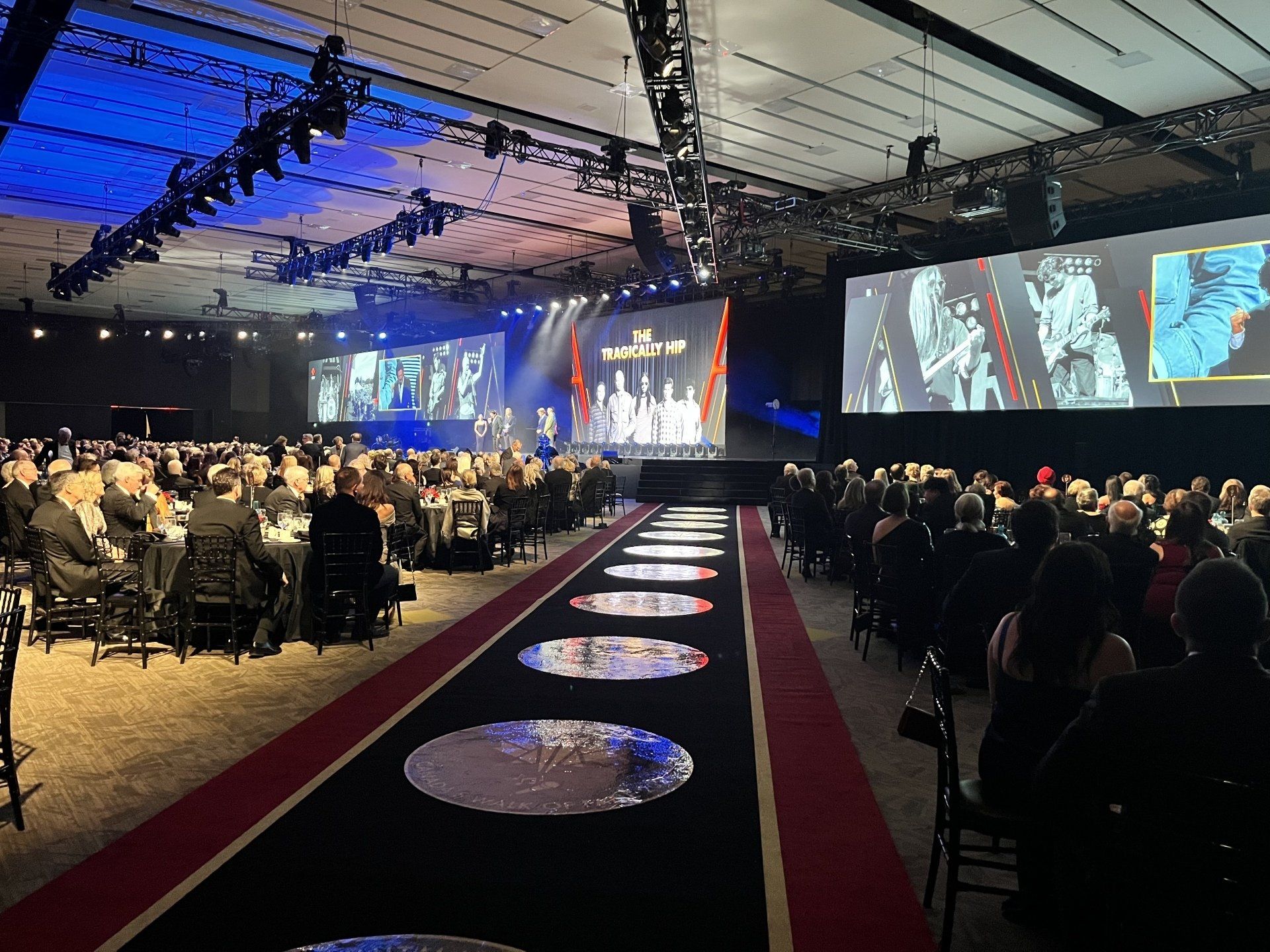 A large group of people are sitting at tables in a large auditorium watching a presentation.